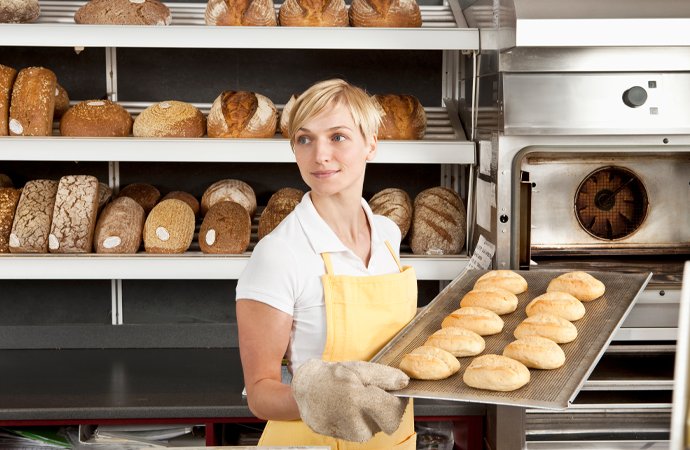 Blonde B&auml;ckerin mit gelber Sch&uuml;rze und wei&szlig;em Poloshirt h&auml;lt ein Backblech mit frisch gebackenen Br&ouml;tchen vor einem ge&ouml;ffneten Backofen. Hinter ihr befinden sich Metallregale mit verschiedenen Brotsorten.