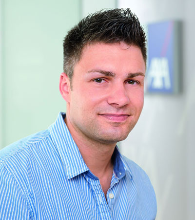 Junger Mann mit kurzen dunklen Haaren und blauer Streifenbluse blickt l&auml;chelnd in die Kamera, im Hintergrund verschwommen das AXA-Logo.