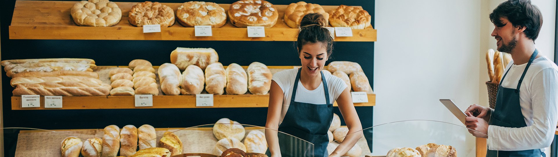 Zwei Mitarbeiter einer B&auml;ckerei stehen hinter einer Verkaufstheke. Die Frau tr&auml;gt eine dunkle Sch&uuml;rze und bereitet Waren vor, w&auml;hrend ihr Kollege ein Tablet bedient. Im Hintergrund stehen Holzregale mit verschiedenen Broten und Backwaren.