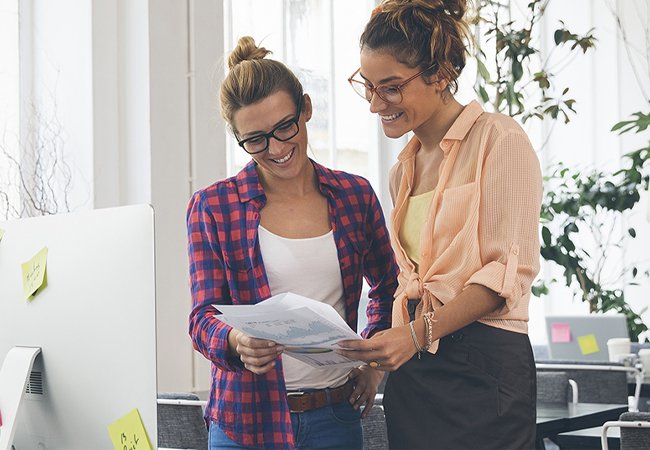 Zwei junge Frauen stehen in einem modernen B&uuml;ro nebeneinander und betrachten gemeinsam ein Dokument. Beide lachen und wirken konzentriert. Auf dem Schreibtisch kleben bunte Notizzettel.
