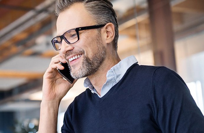 Ein lächelnder Mann mit Brille und Bart telefoniert mit seinem Smartphone. Er trägt ein dunkelblaues Oberteil über einem hellblauen Hemd und wirkt entspannt.