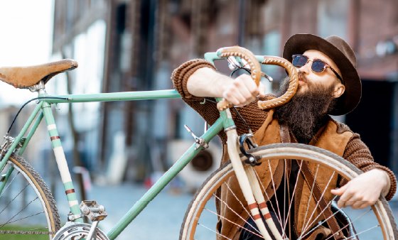 Ein Mann mit Vollbart, Sonnenbrille, Hut und hellbrauner Jacke umarmt sein hellblaues Rennrad.