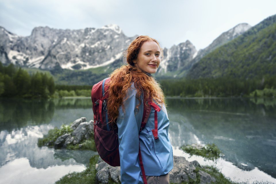 Junge Frau mit rotem Rucksack steht an einem Bergsee und blickt &uuml;ber die Schulter, im Hintergrund spiegeln sich bewaldete Berge im Wasser.