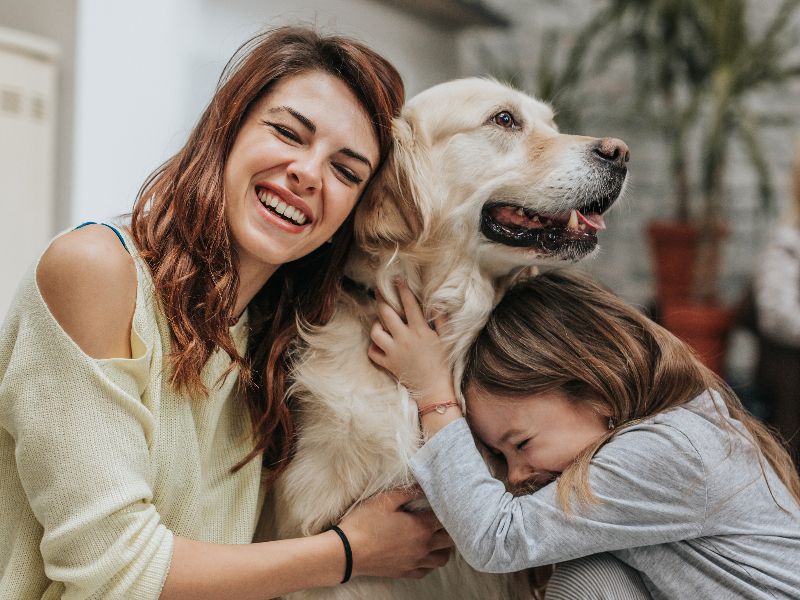 Tierhalterhaftpflichtversicherung | AXA Frau und Kind umarmen einen Golden Retriever in einem Wohnzimmer. Alle drei wirken fröhlich und verbunden, die Frau lacht in die Kamera.