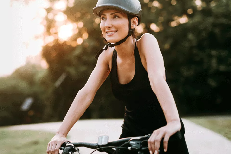 Eine junge Frau mit schwarzem Helm fährt lächelnd auf einem Fahrrad durch einen Park. 
