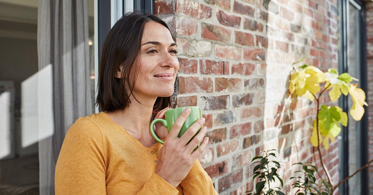 Eine Frau steht entspannt vor einer Ziegelwand und h&auml;lt eine gr&uuml;ne Tasse in beiden H&auml;nden. Sie blickt l&auml;chelnd in die Ferne.