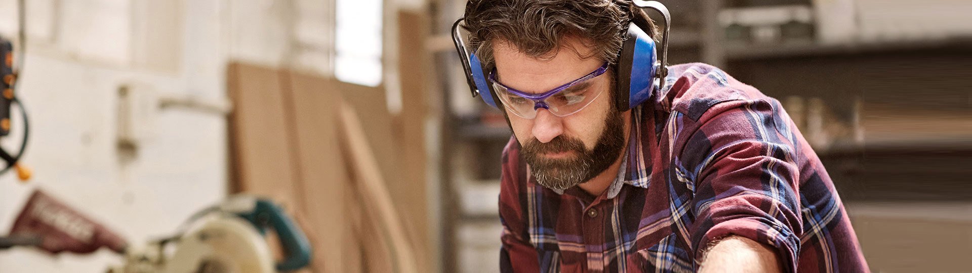 Ein bärtiger Mann in kariertem Holzfällerhemd arbeitet konzentriert in einer Werkstatt. Er trägt Gehörschutz und Schutzbrille und beugt sich über eine Werkbank.