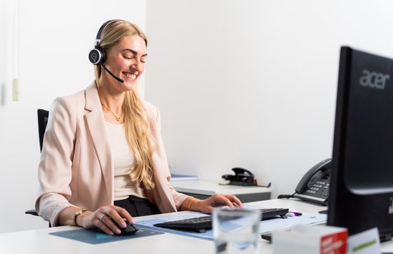 Kundenservice im Büro mit Headset Eine junge Frau mit langem blondem Haar sitzt lächelnd an einem Schreibtisch im Büro. Sie trägt ein Headset und bedient Maus und Tastatur. Vor ihr steht ein Monitor, auf dem Schreibtisch befinden sich ein Glas Wasser, ein Telefon und Büromaterialien.