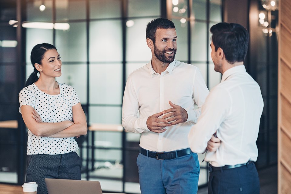 Drei Geschäftsleute stehen im modernen Büro zusammen und führen ein Gespräch. Ein Mann erklärt etwas gestikulierend, während eine Frau und ein weiterer Mann aufmerksam zuhören.