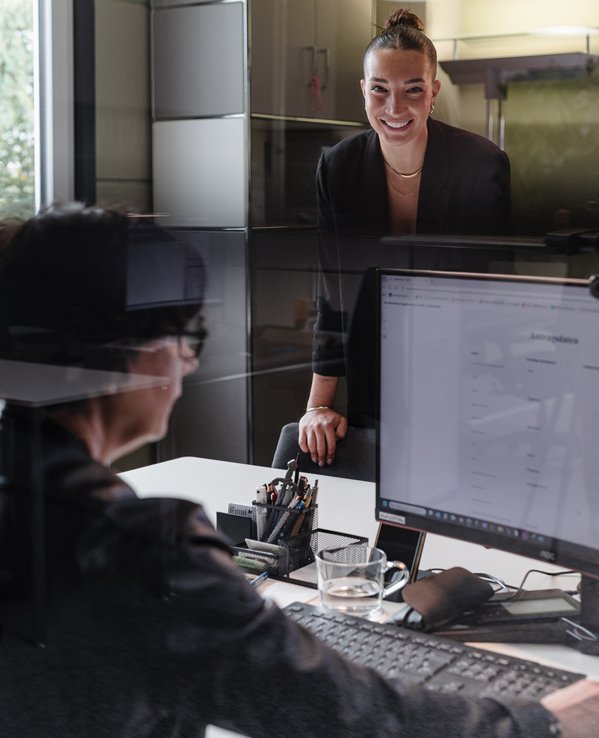 Eine Beraterin lehnt l&auml;chelnd an einem Schreibtisch in einem modernen B&uuml;ro und spricht mit einer Kundin, die vor einem Computerbildschirm sitzt.