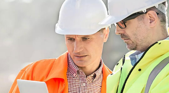Zwei Bauarbeiter mit Schutzhelmen und Warnkleidung stehen im Freien und schauen gemeinsam auf ein Tablet. 
