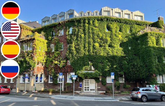 Unsere Beratersprachen | AXA Versicherung Joepen & Schmid oHG in Mönchengladbach Außenansicht eines Bürogebäudes mit vollständig bewachsener Fassade aus grünem Efeu. Vor dem Gebäude stehen mehrere Autos und Parkschilder. Am linken Bildrand sind vier runde Flaggen zu sehen: Deutschland, USA, Spanien und Russland.