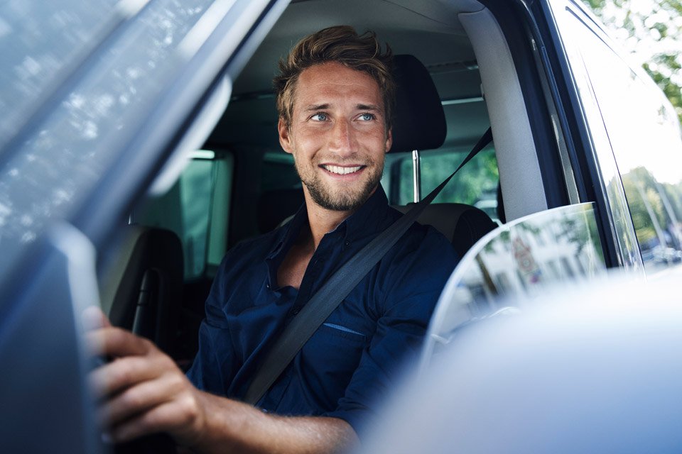 Ein junger Mann mit Bart und dunkelblauem Hemd sitzt angeschnallt am Steuer eines Autos und lächelt nach draußen. Die Perspektive zeigt ihn durch das offene Seitenfenster.