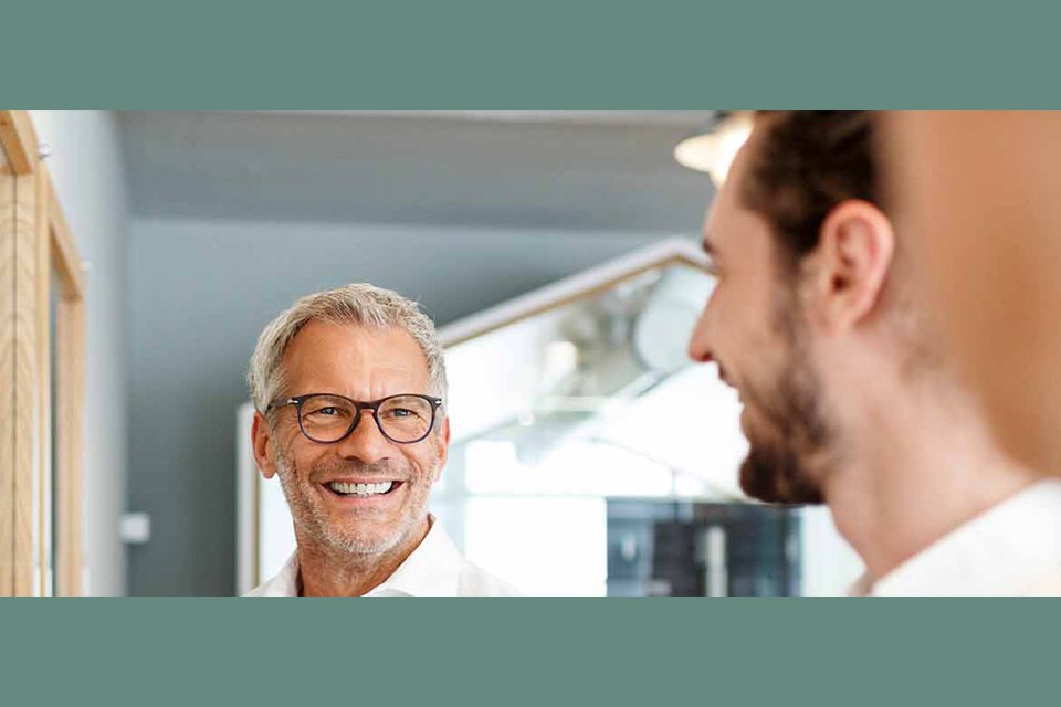 Zwei Männer unterhalten sich lächelnd in einem modernen Büroflur. Im Vordergrund ist ein älterer Mann mit Brille zu sehen, der freundlich den anderen ansieht.