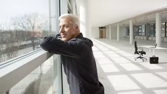&Auml;lterer Mann mit kurzen wei&szlig;en Haaren und dunklem Pullover lehnt an einem Gel&auml;nder vor einer gro&szlig;en Fensterfront. Er blickt l&auml;chelnd nach drau&szlig;en. Hinter ihm erstreckt sich ein heller, weitl&auml;ufiger B&uuml;roraum.