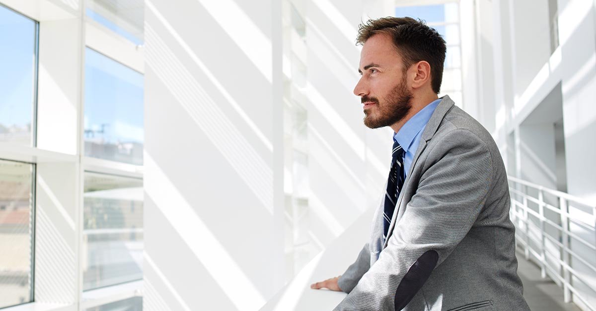 Ein junger Mann im grauen Anzug und gestreifter Krawatte blickt nachdenklich zur Seite. Er steht in einem lichtdurchfluteten, modernen B&uuml;ro mit wei&szlig;en W&auml;nden und gro&szlig;en Fenstern.