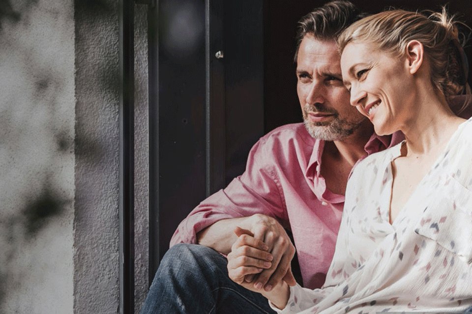 Ein Paar blickt entspannt aus dem Fenster ihres Hauses. Der Mann trägt ein rosa Hemd, die Frau lehnt sich lächelnd an ihn. Die Szene vermittelt Geborgenheit und Vertrauen.