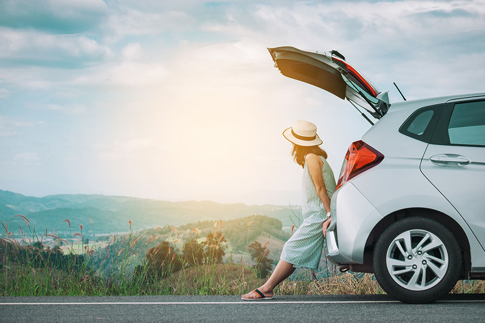 Frau in Sommerkleid und Strohhut lehnt entspannt am offenen Kofferraum eines silbernen Autos, geparkt an einer Landstraße mit Blick auf grüne Hügel und weite Landschaft im Sonnenlicht.