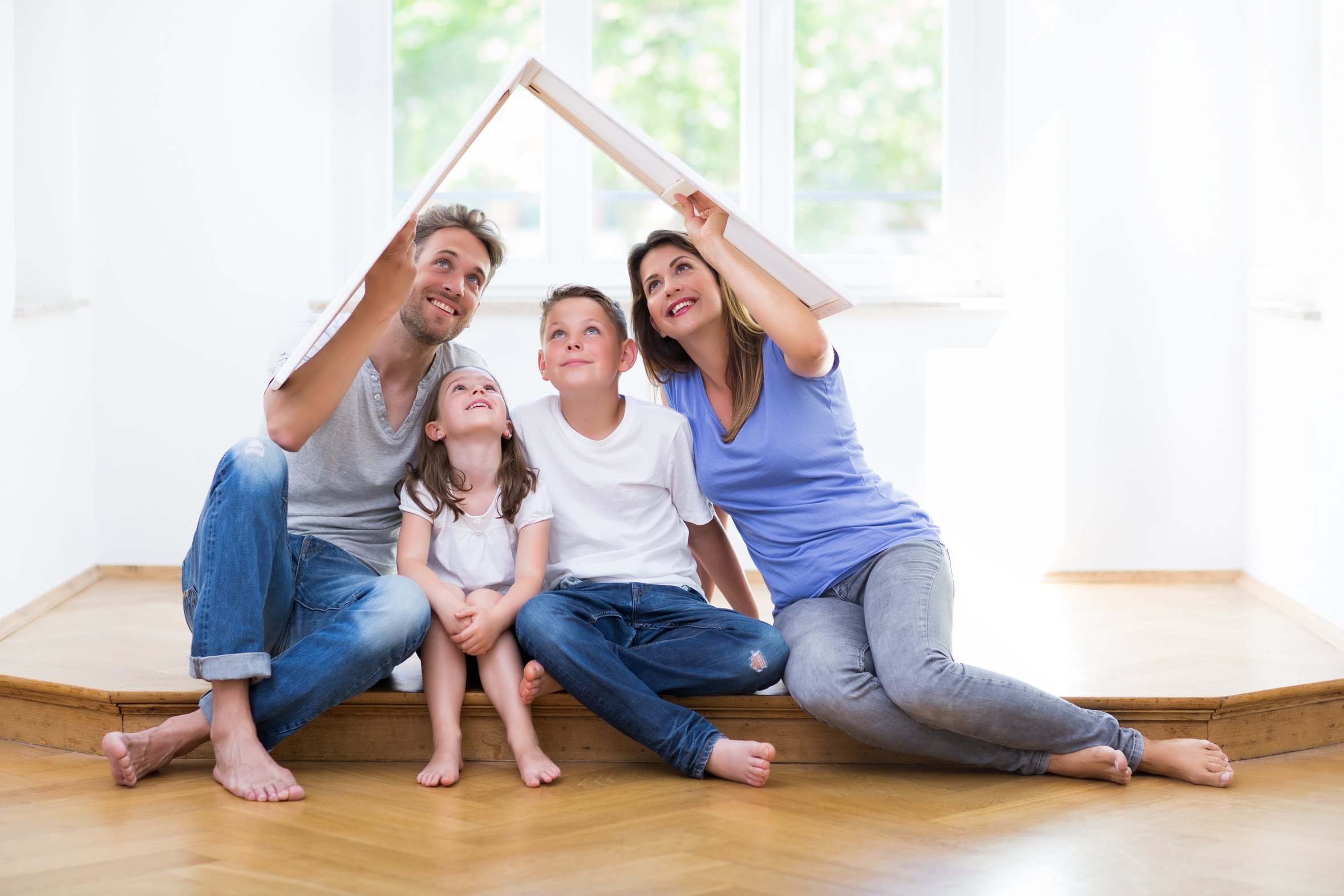 Eine vierk&ouml;pfige Familie sitzt barfu&szlig; auf dem Boden eines hellen, leeren Wohnraums. Zwei Erwachsene und zwei Kinder halte