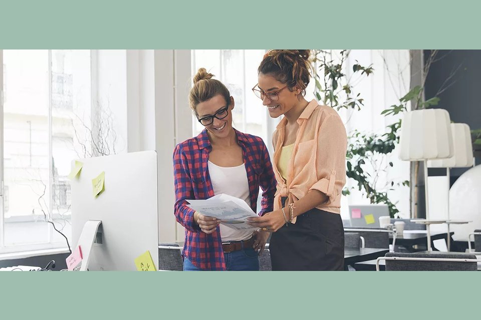 Zwei junge Frauen stehen in einem modernen Büro vor einem Schreibtisch und betrachten gemeinsam ein Dokument. Sie lachen und wirken zufrieden. Der Arbeitsplatz ist mit Post-its, Pflanzen und Designmöbeln gestaltet.