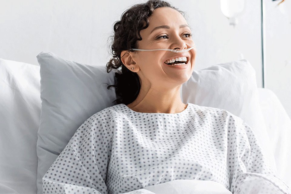  Ein Mann mit blauem Tshirt liegt in einem Krankenbett und l&auml;chelt eine blonde Frau mit rosa Oberteil an, weil er umsorgt wird.