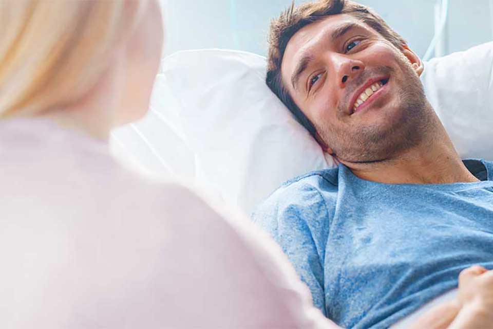 Ein lächelnder Mann liegt in einem Krankenhausbett und blickt eine Frau an, die an seiner Seite sitzt.