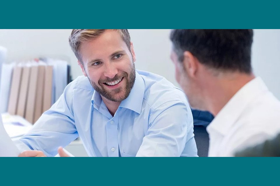 Zwei Männer im Gespräch in einem modernen Büro. Der Mann im Vordergrund lächelt freundlich und trägt ein hellblaues Hemd, während der andere im Profil zu sehen ist.