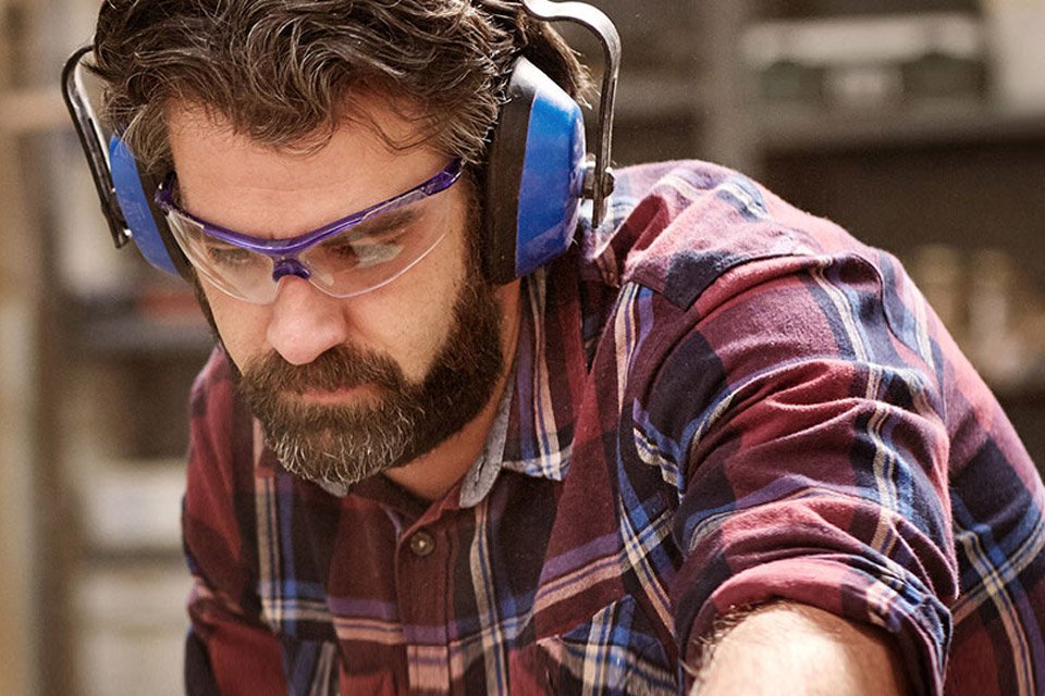 Ein Mann mit Bart trägt ein kariertes Hemd, Gehörschutz und Schutzbrille. Er arbeitet konzentriert an einem Werkstück in einer Holzwerkstatt.