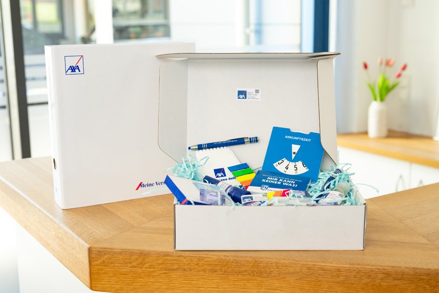 Offene Willkommensbox mit AXA-Logo, gefüllt mit Schreibwaren, Informationsmaterial und kleinen Geschenken, auf einer Holztheke platziert.