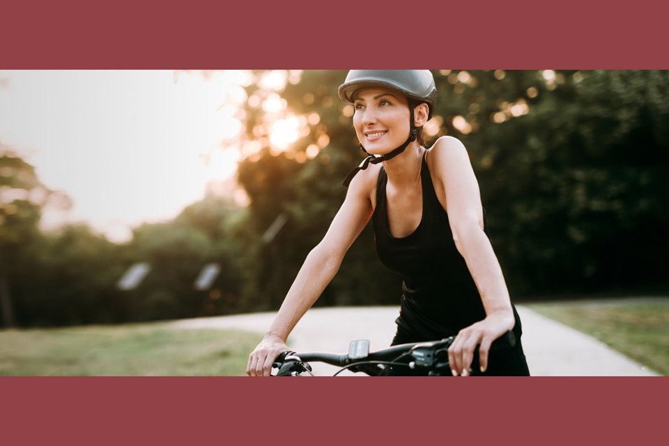 Junge Frau mit Fahrradhelm fährt bei Sonnenuntergang lächelnd auf einem Weg durch einen Park.