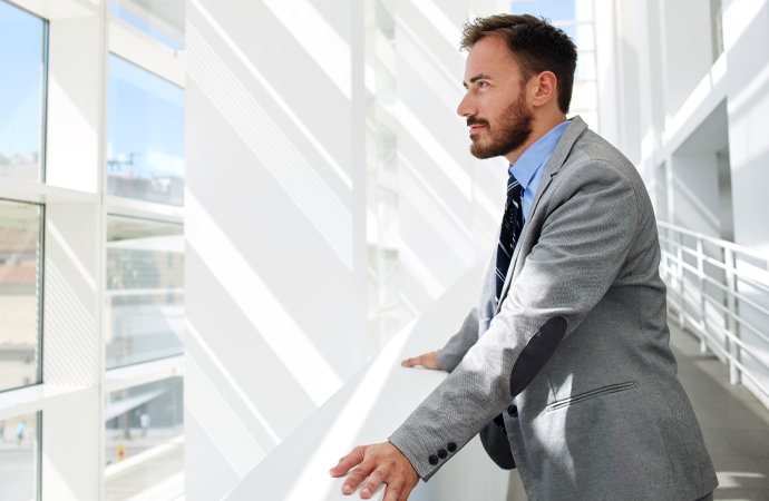 Ein junger Mann im grauen Anzug und gestreifter Krawatte blickt nachdenklich zur Seite. Er steht in einem lichtdurchfluteten, modernen B&uuml;ro mit wei&szlig;en W&auml;nden und gro&szlig;en Fenstern.