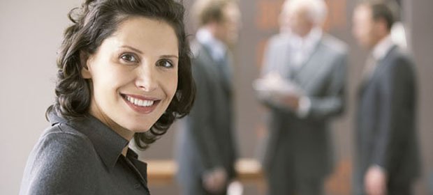 L&auml;chelnde Gesch&auml;ftsfrau im Vordergrund eines B&uuml;ros, im Hintergrund sprechen drei Anzugtr&auml;ger &uuml;ber Dokumente.