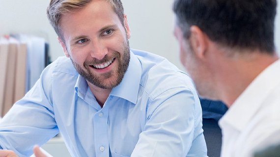 Zwei M&auml;nner im B&uuml;ro im Gespr&auml;ch. Einer von ihnen h&auml;lt ein Dokument in der Hand und l&auml;chelt seinem Gegen&uuml;ber zu. Im Hintergrund stehen Aktenordner und Computerbildschirme.