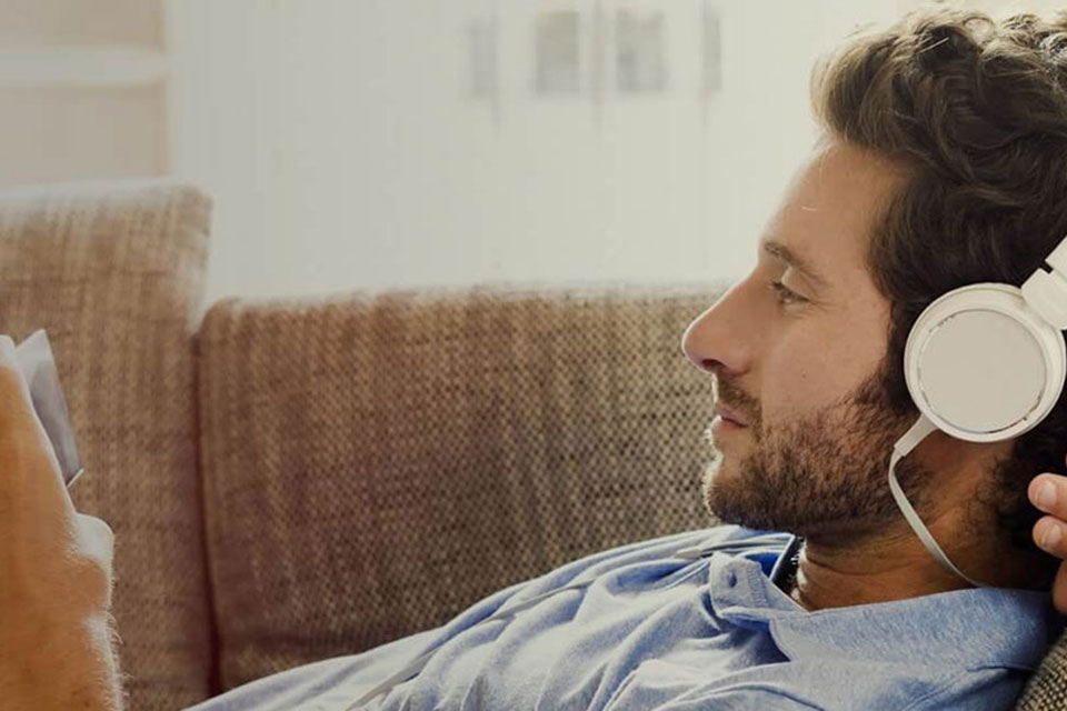 Ein Mann im blauen Hemd liegt auf dem Sofa und schaut auf sein Smartphone, dabei hat er wei&szlig;e Kopfh&ouml;rer auf.
