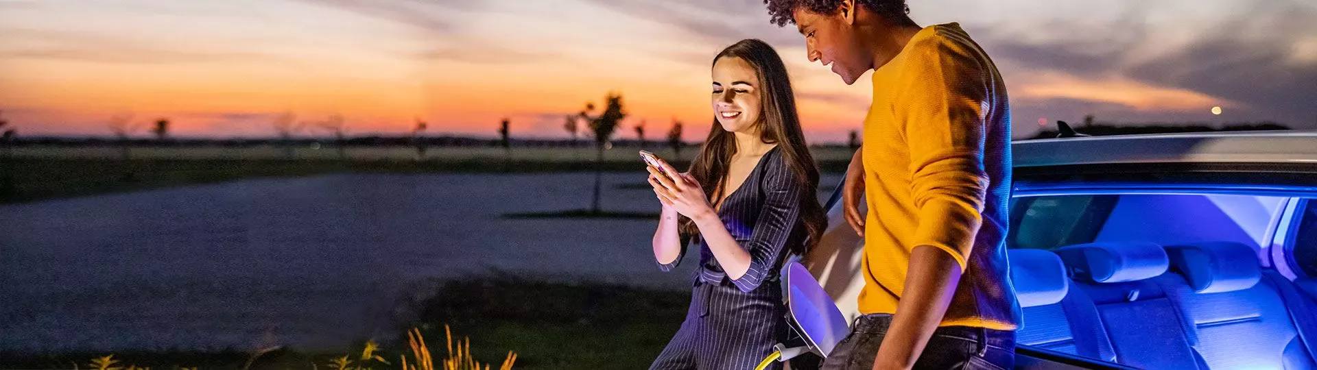 Ein junges Paar steht neben einem Elektroauto bei Sonnenuntergang, die Frau h&auml;lt ein Smartphone in der Hand, beide l&auml;cheln. Im Hintergrund eine offene Landschaft mit orangenem Abendhimmel.