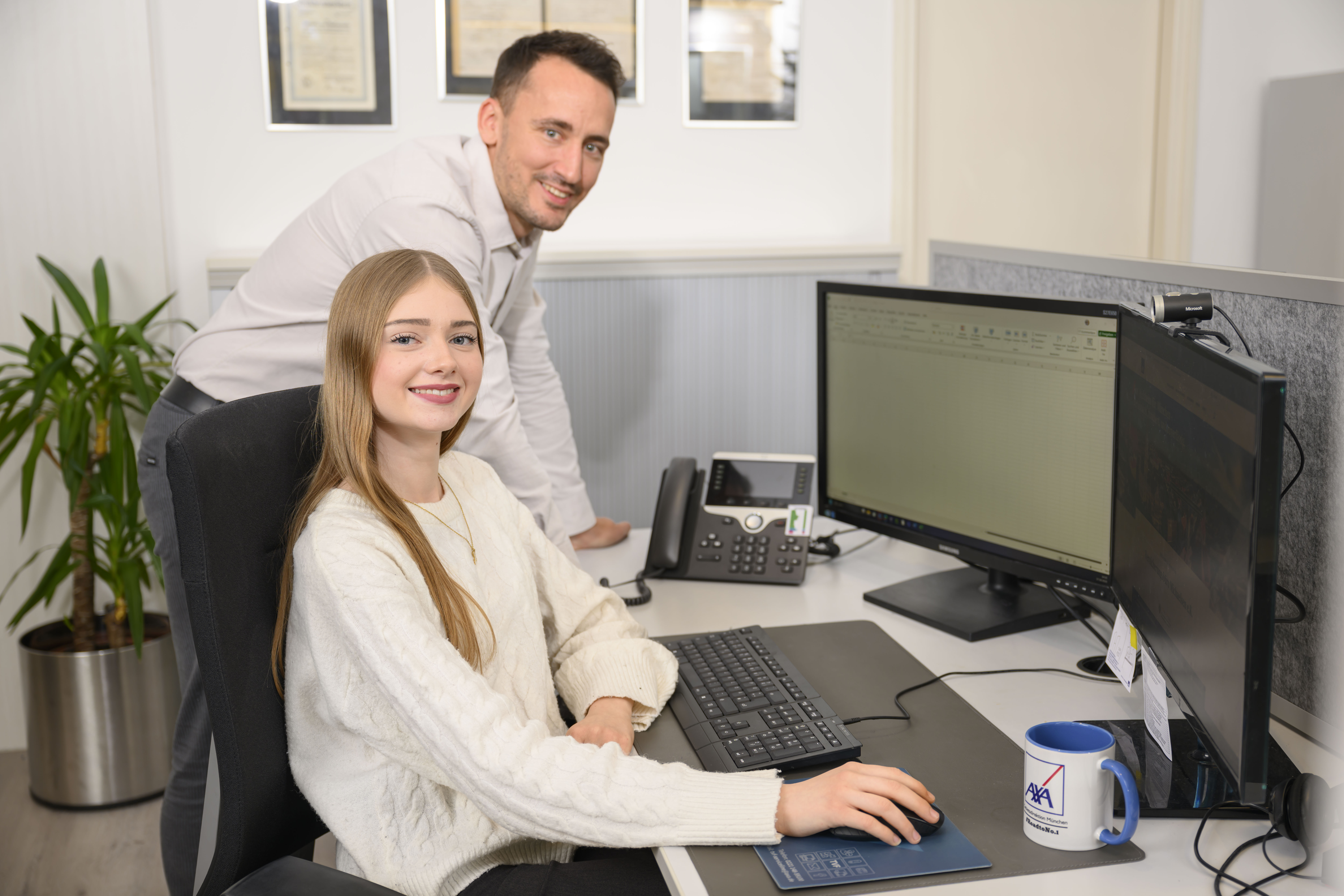 Junge Mitarbeiterin am Schreibtisch mit Tastatur und zwei Monitoren, daneben ein Kollege, beide lächelnd in Richtung Kamera.