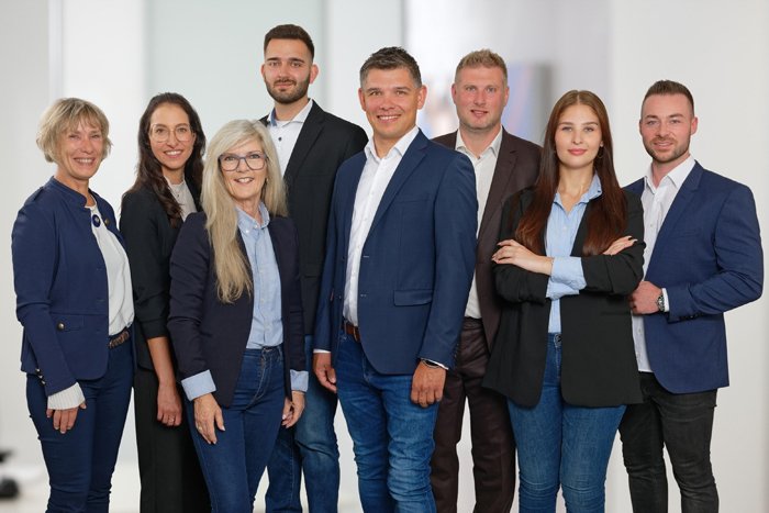  Acht Personen stehen in einem hellen Büro und lächeln in die Kamera. Sie tragen Businesskleidung mit Blazern, Hemden und Jeans, wirken professionell und freundlich. Zwei Frauen stehen mit verschränkten Armen, die Gruppe wirkt als Team geschlossen und sympathisch.