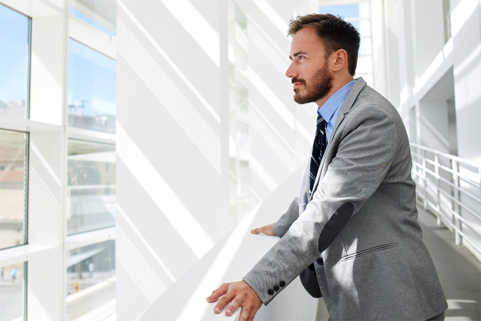 Ein junger Mann im grauen Anzug und gestreifter Krawatte blickt nachdenklich zur Seite. Er steht in einem lichtdurchfluteten, modernen B&uuml;ro mit wei&szlig;en W&auml;nden und gro&szlig;en Fenstern.
