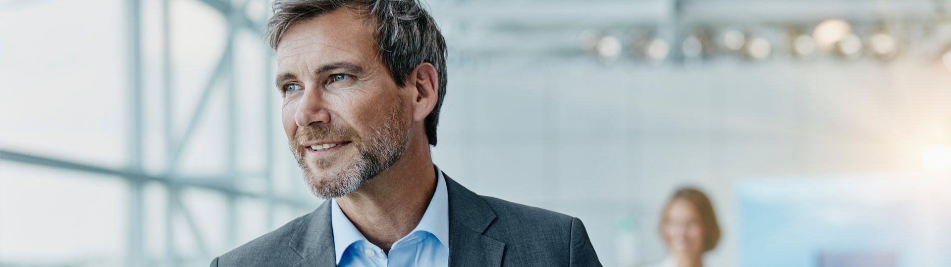 Gesch&auml;ftsmann mit kurzen grauen Haaren, Bart und grauem Sakko steht in einem hellen B&uuml;rogeb&auml;ude mit gro&szlig;en Glasfenstern. Er l&auml;chelt und blickt nachdenklich zur Seite.
